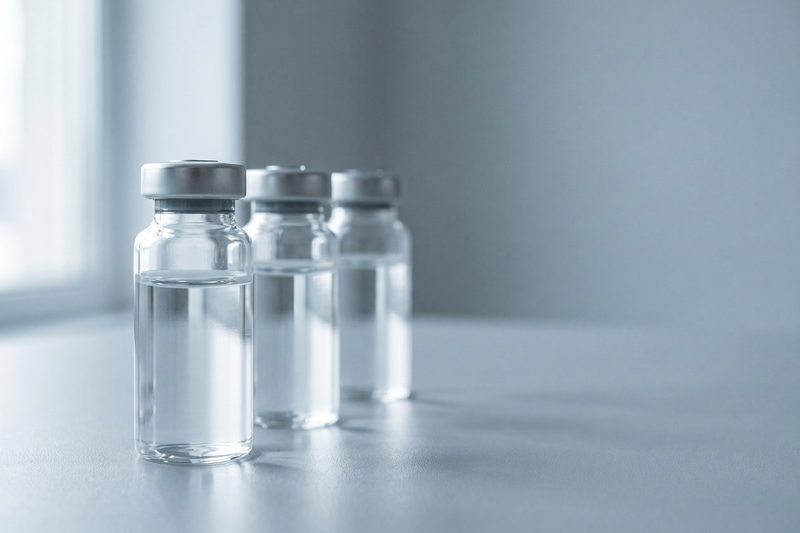 Drei unbeschriftete Glasfläschchen mit klarer Lösung auf hellgrauem Studiogrund.